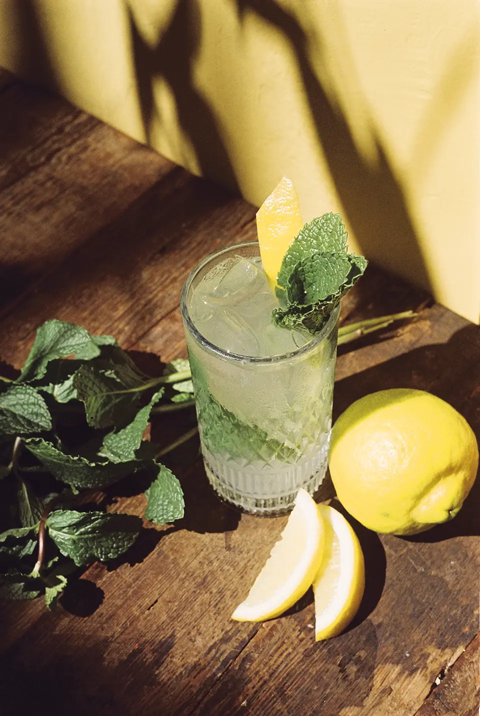 Um copo sobre uma superfície de madeira escura. O copo está cheio de um drink verde bem claro, com gelo, folhas de hotelã e um zest de limão siliano. Ao redor do copo há ramos de hortelã e um limão siciliano.