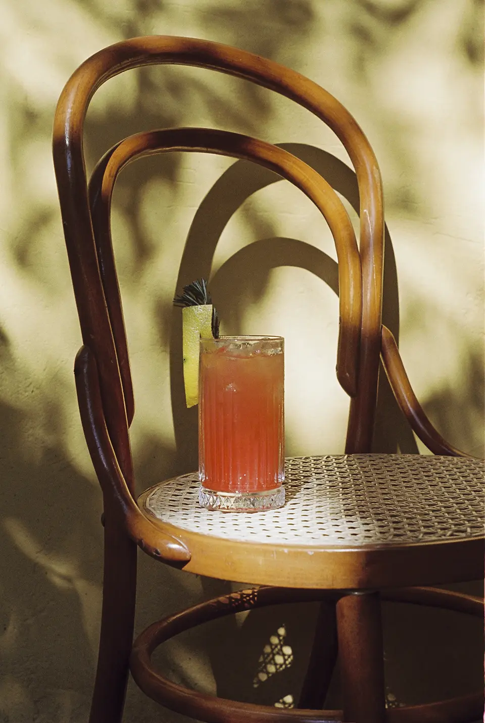 Um copo sobre uma cadeira de madeira cor de caramelo, com assento trançado. O copo está cheio de um drink avermelhado com gelo, um zest de limão siciliano e um ramo de alecrim.