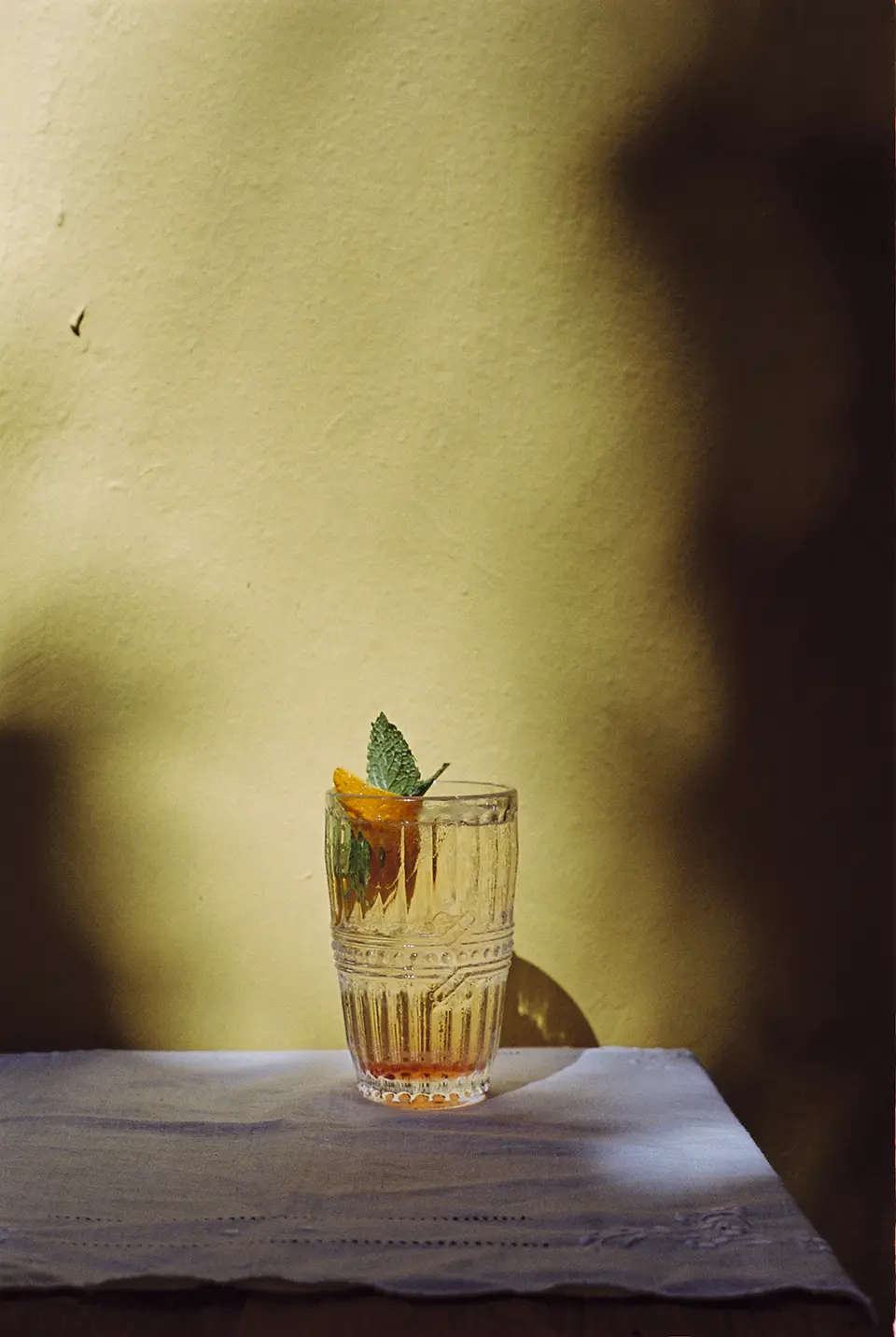 Um copo com um drink transparente, levemente alaranjado repousa sobre uma mesa coberta com uma tolha branca. Dentro do copo há também meia fatia de limão siciliano e um ramo de hortelã.